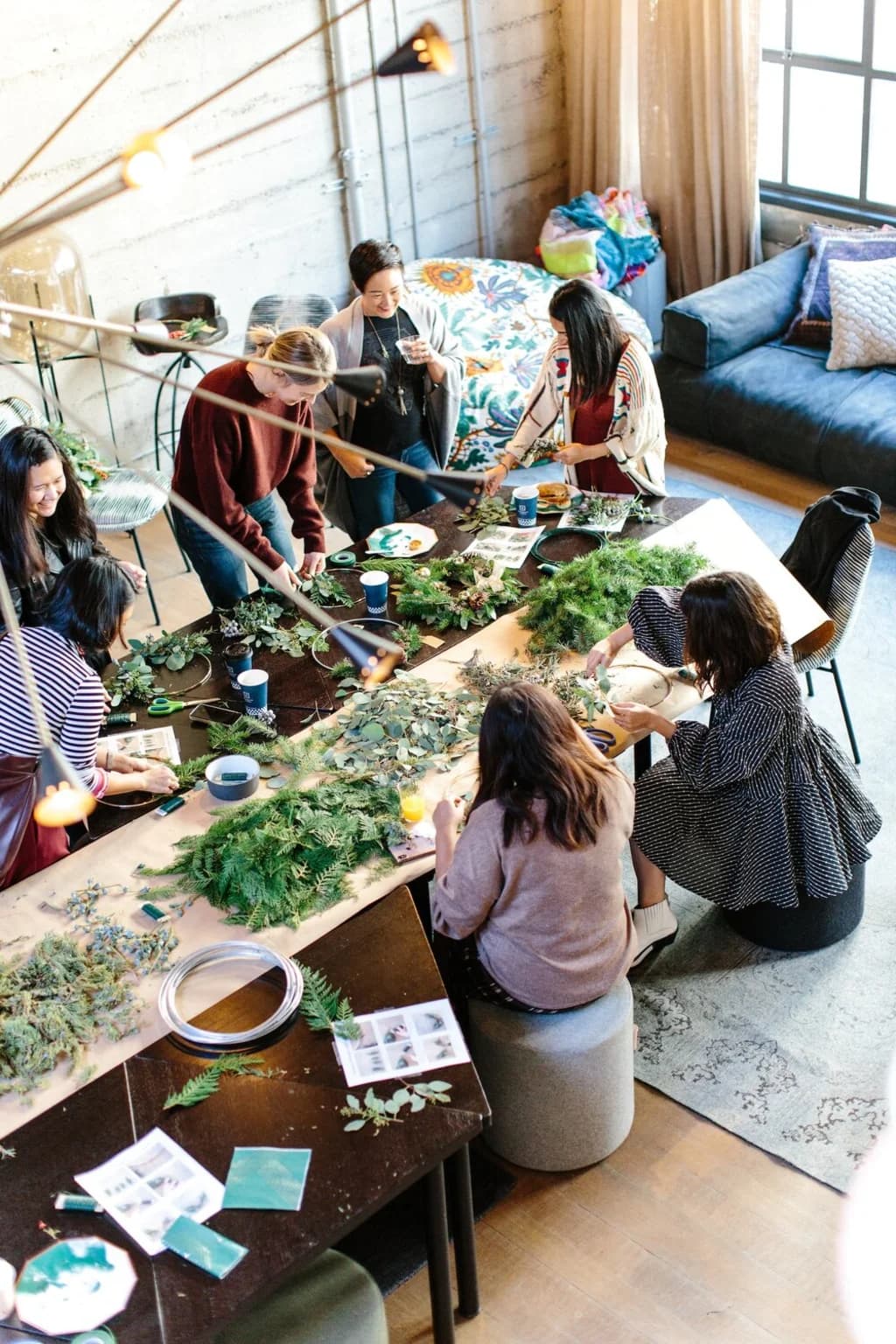 Overhead view of a group of people gathered around a large table, working together on crafting holiday wreaths with various types of greenery and decorations.